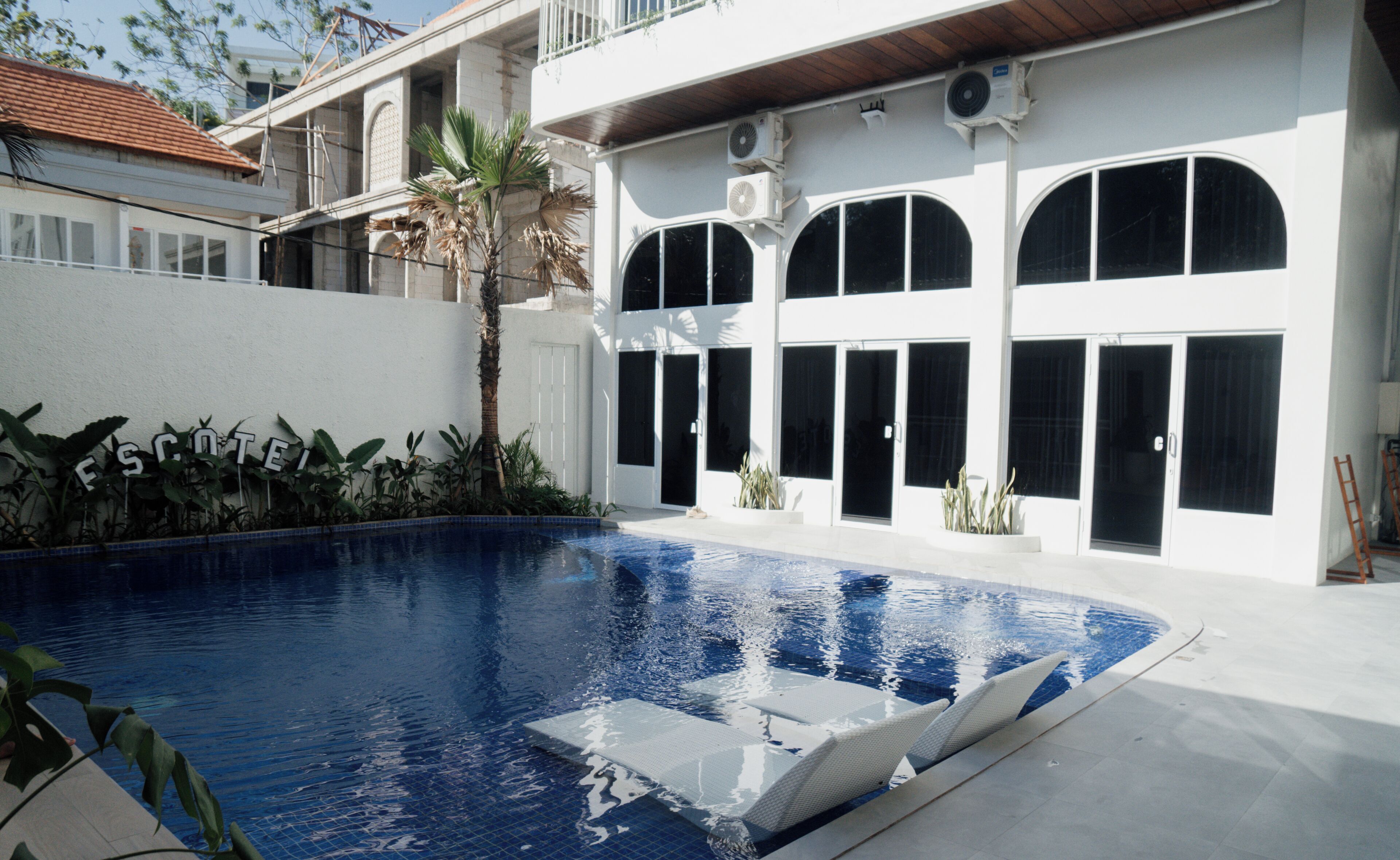 A swimming pool with a white deck, surrounded by a white building with large arched windows and a palm tree.