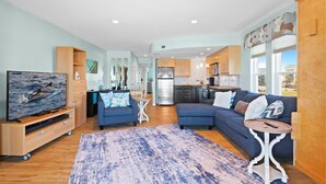 A modern living room with a large TV, blue sofas, and a kitchen in the background. - The Endless Wave: Oceanfront Condo, Sunrise View, Pool, 1-mile to Dining & Pier (Surf City)