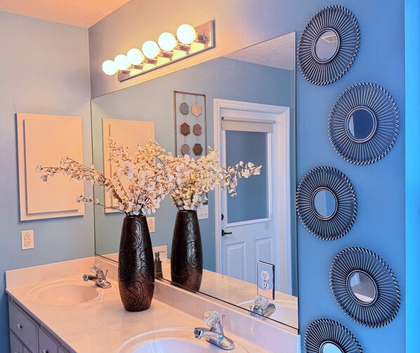 A bathroom with two dark vases with floral arrangements, a mirror with lights above, and decorative sunburst mirrors on the wall. - Peaceful, Relaxing & Quiet Home With A Private Fenced Yard In Ocala FL (Ocala)