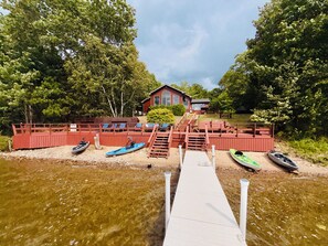Un muelle de madera que lleva a una casa con fachada roja, rodeada de árboles y una masa de agua tranquila.
