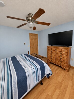 A bedroom with a bed, a wooden dresser, a ceiling fan, a flat-screen TV, and a door.