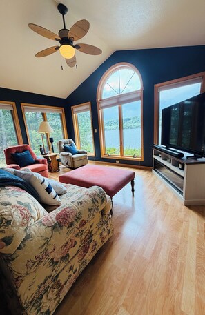 A room with a large bed, a television, and a view of a lake through large windows. - Fall Lakefront Escape on Monocle Lake with Private Dock + Bunkhouse (Brimley)