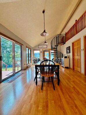 A spacious dining area with a wooden table and chairs, a spiral staircase, and large windows offering a view of the outdoors. - Fall Lakefront Escape on Monocle Lake with Private Dock + Bunkhouse (Brimley)