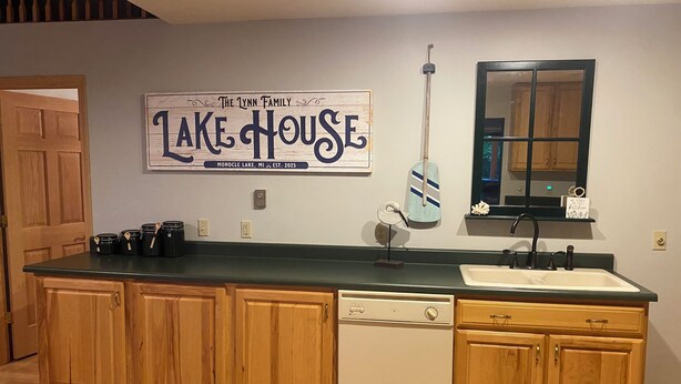Eine Küche mit Holzausstattung, einem Geschirrspüler, einer Spüle und einem Schild mit der Aufschrift 'The Lynn Family Lake House Monocle Lake, MI Established 2025'.