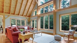 Una habitación amplia con techo alto, ventanas grandes, un sofá rojo, una mesa de centro de madera y un sillón mecedora.