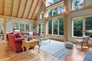 Une pièce spacieuse avec un plafond haut, de grandes fenêtres, un canapé rouge, une table basse en bois et un fauteuil à bascule.