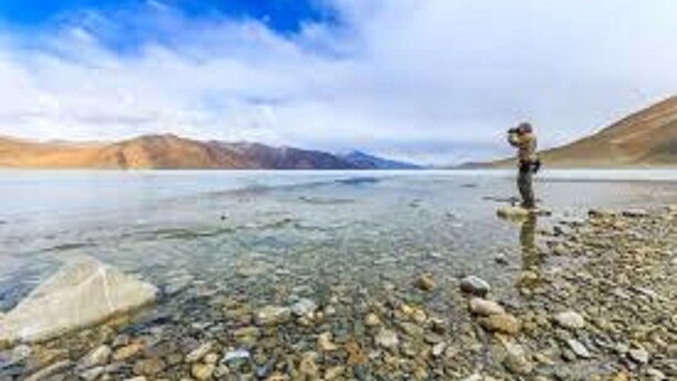 Une personne se tient sur une côte rocheuse, au bord d’un lac paisible, avec des montagnes en arrière-plan.