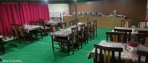 A restaurant with multiple wooden tables and chairs, a green carpeted floor, and a counter with kitchen equipment in the background. - Himalayan Inn Cottages - Pangong Lake (Leh)