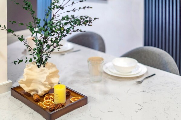 Une table à manger recouverte d’une nappe blanche, un vase décoratif avec des feuilles vertes, une bougie et un plateau contenant des tranches d’orange séchées et des noix.