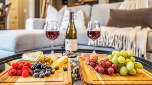 A dining table with a variety of cheeses, fruits, and a bottle of wine, set in a modern living room with a sofa and a bookshelf in the background. - Eagle-Vail Escape | 3BR Near Skiing & Trails (Vail)