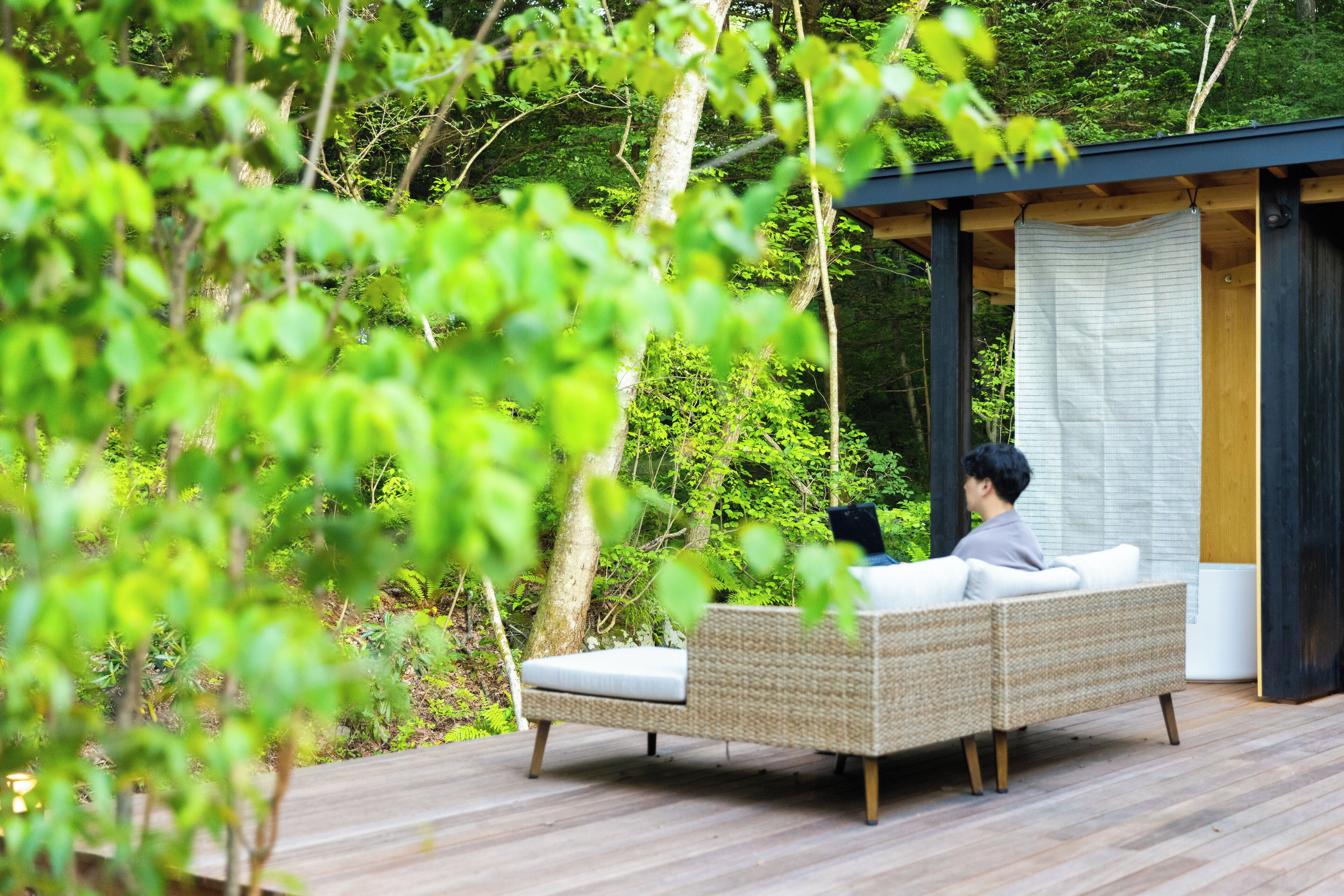 A wooden deck with a wicker sofa and a small table, surrounded by lush greenery.