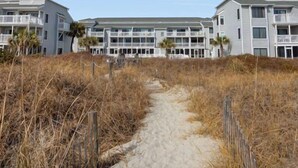 A beachfront hotel with multiple-story buildings, a sandy pathway leading to the entrance, and a clear blue sky. - Sea Cloisters 105b (North Myrtle Beach)
