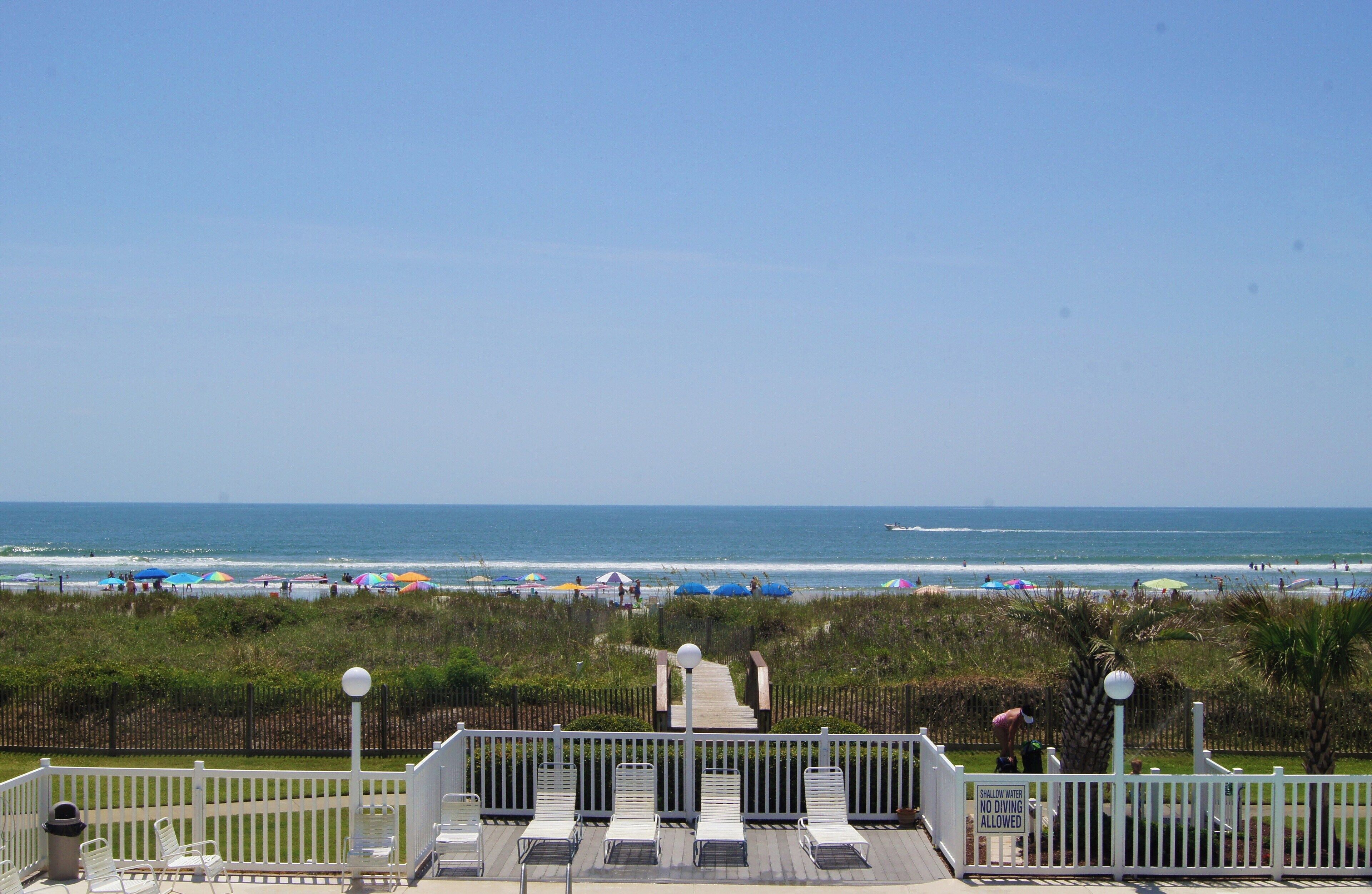 A beach view with a wooden deck, white railings, and a sign indicating shallow water no diving allowed.