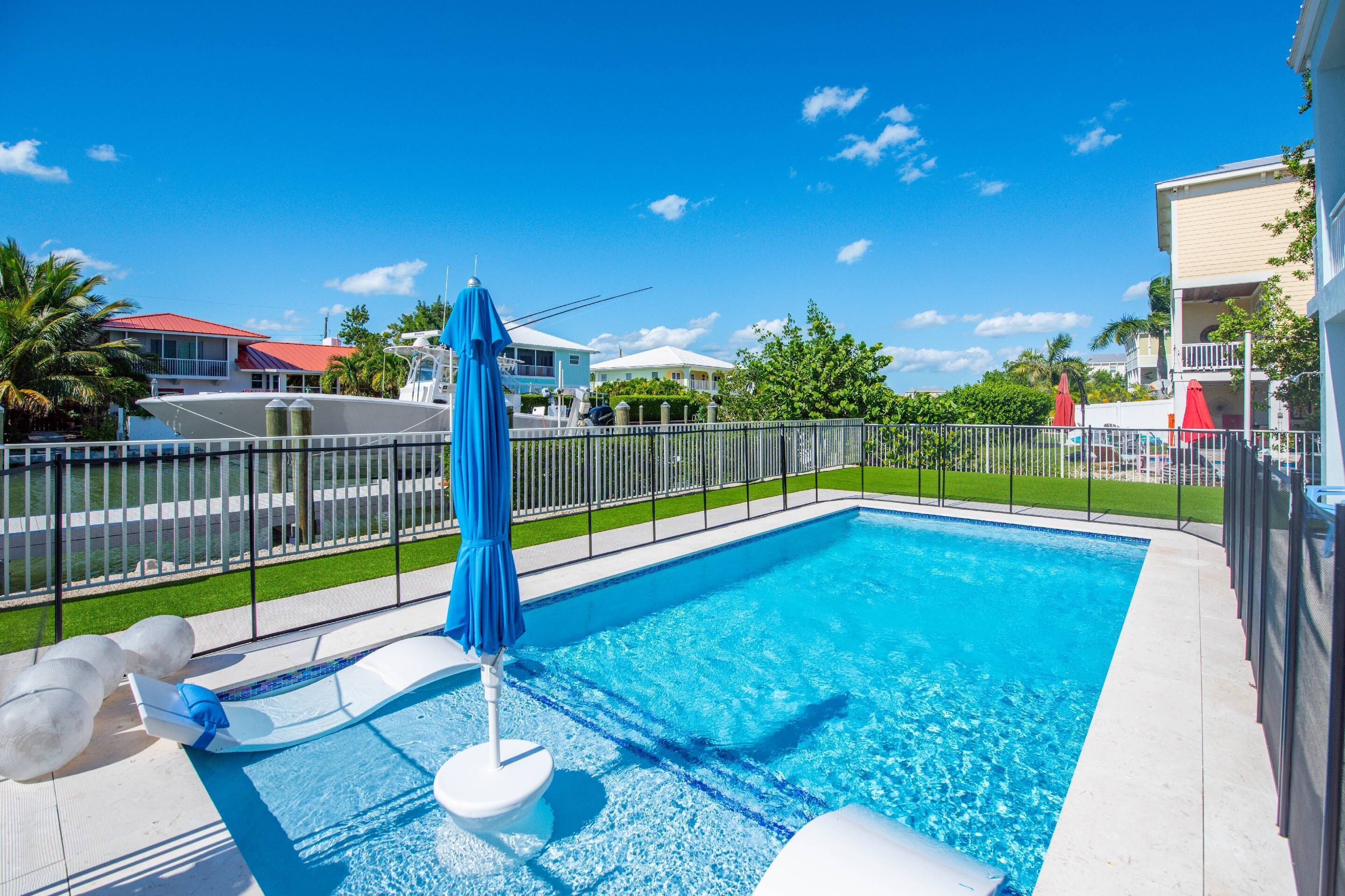 Una piscina con sombrilla azul, sillas de descanso y una zona vallada con casas al fondo.