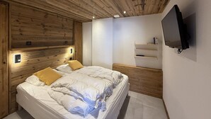 Un dormitorio con techo de madera, una cama con ropa de cama blanca y una almohada amarilla, un televisor montado y un pequeño estante.