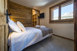 Une chambre à coucher avec une tête de lit en bois, un lit recouvert de draps blancs, un téléviseur fixé au mur et une fenêtre donnant sur les montagnes.