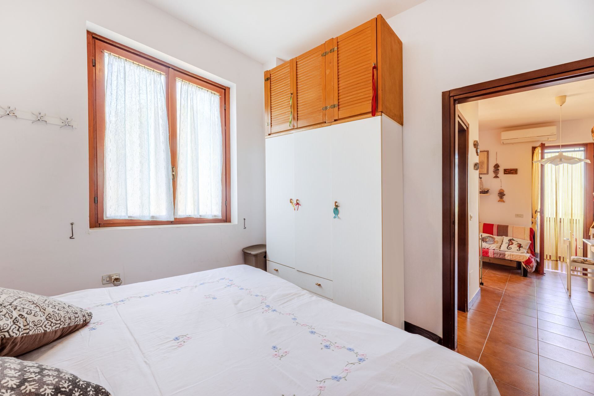 A bedroom with a bed, a wardrobe, a window with curtains, and a door leading to another room.