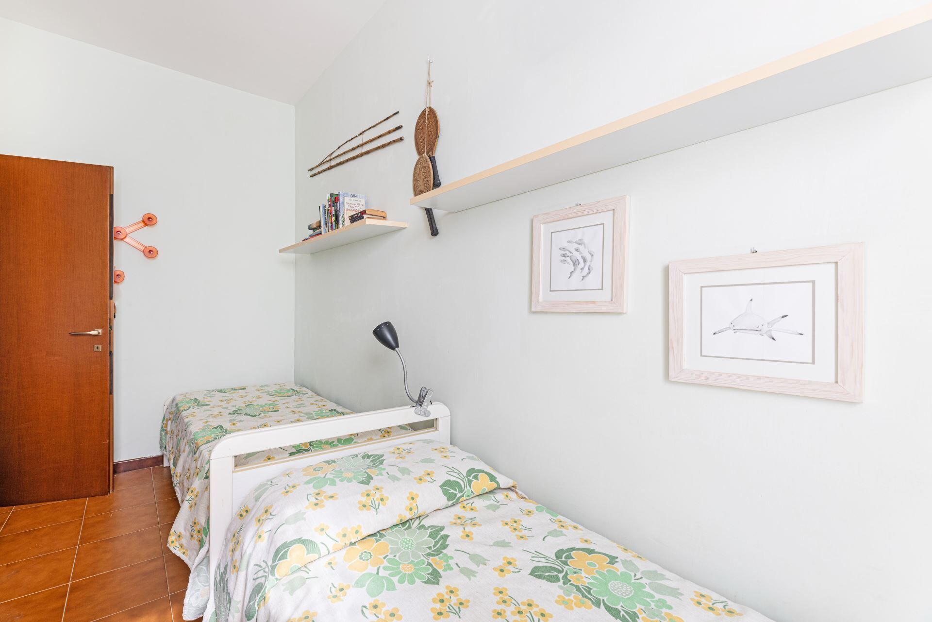 Une chambre avec deux lits simples, une porte en bois, une étagère avec des livres et des objets de décoration, et deux tableaux encadrés accrochés au mur.