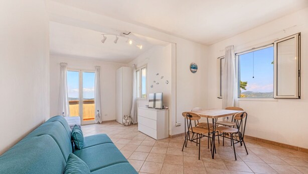 A living room with a teal sofa, a dining table with chairs, a TV on a white cabinet, and a large window with a view.