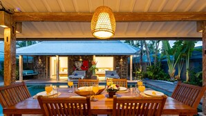 A wooden dining table set for four with a pool and outdoor kitchen in the background. - Villa Yang Tao 4 (Seminyak)