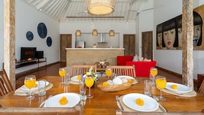 A dining area with a wooden table set for a meal, a kitchen in the background, and a red sofa. - Villa Yang Tao 4 (Seminyak)