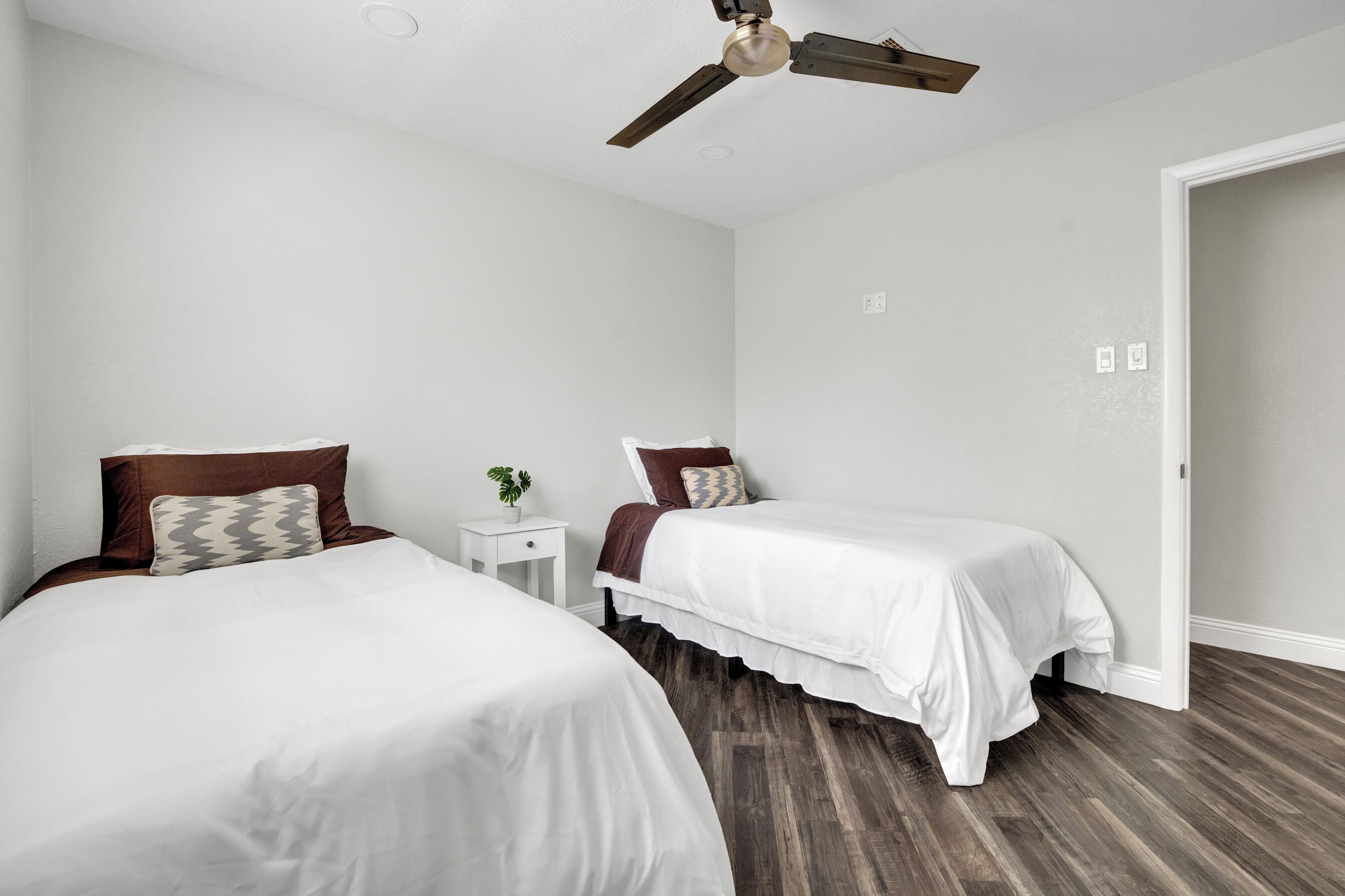 Een hotelkamer met twee eenpersoonsbedden, een plafondventilator, een klein nachtkastje met een plant en een houten vloer.