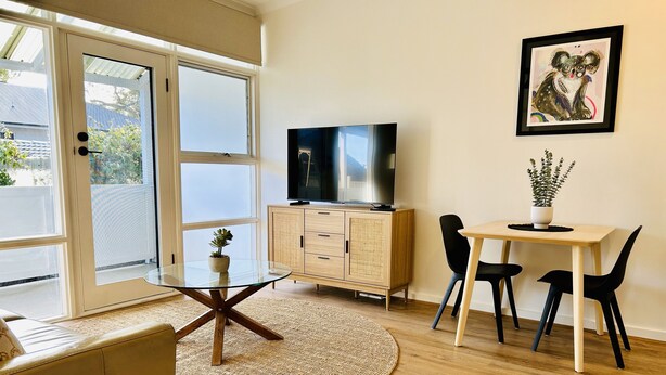 A modern living room with a glass-top coffee table, a sofa, a dining area with a small table and chairs, a television on a wooden cabinet, and a framed artwork on the wall. - Stunning Modern Self-contained Apartment (Hazelwood Park)
