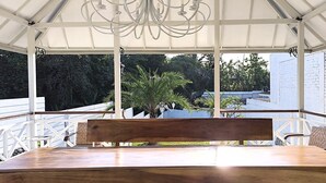 A covered outdoor seating area with a wooden table and bench, white railings, and a view of greenery and a brick wall with decorative plates. - Omah Putih Menoreh (Moyudan)