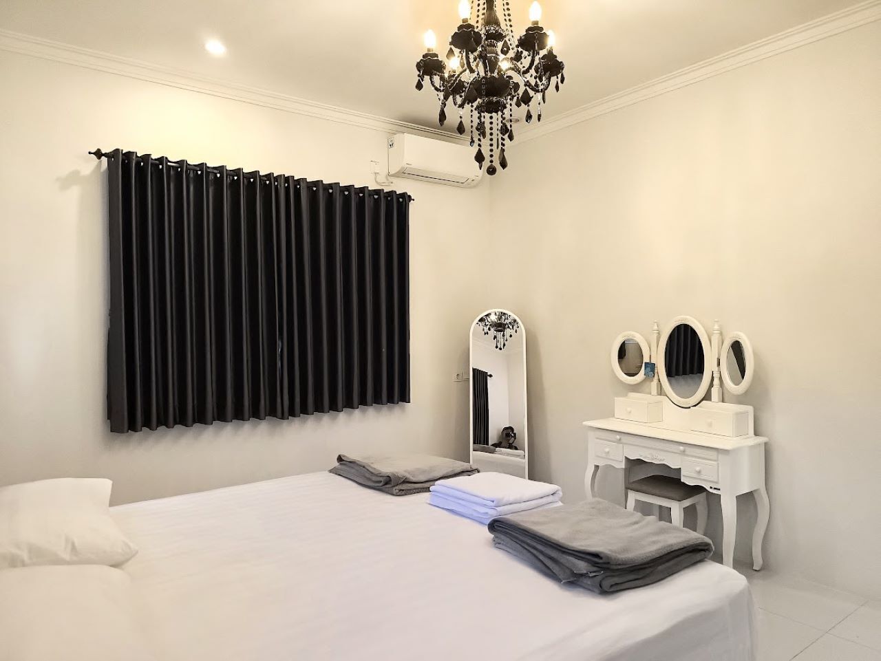 A bedroom with a bed, a vanity with mirrors, a chandelier, and a window with black curtains.