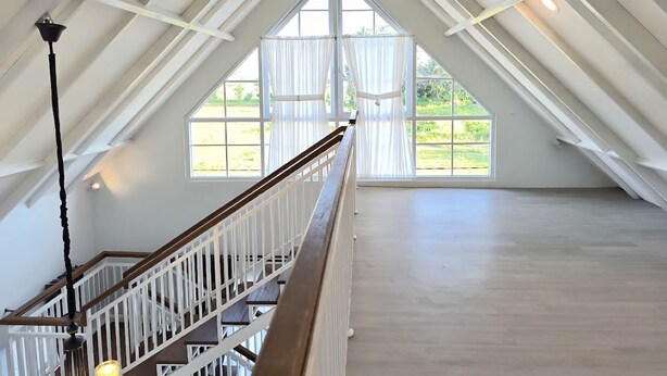 A spacious attic room with a wooden staircase, large windows, and a sloped ceiling. - Omah Putih Menoreh (Moyudan)