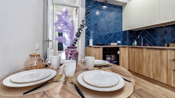 Une cuisine moderne avec un mur recouvert de carreaux bleus, des armoires en bois et un coin repas agrémenté d’une vaisselle blanche et de couverts.