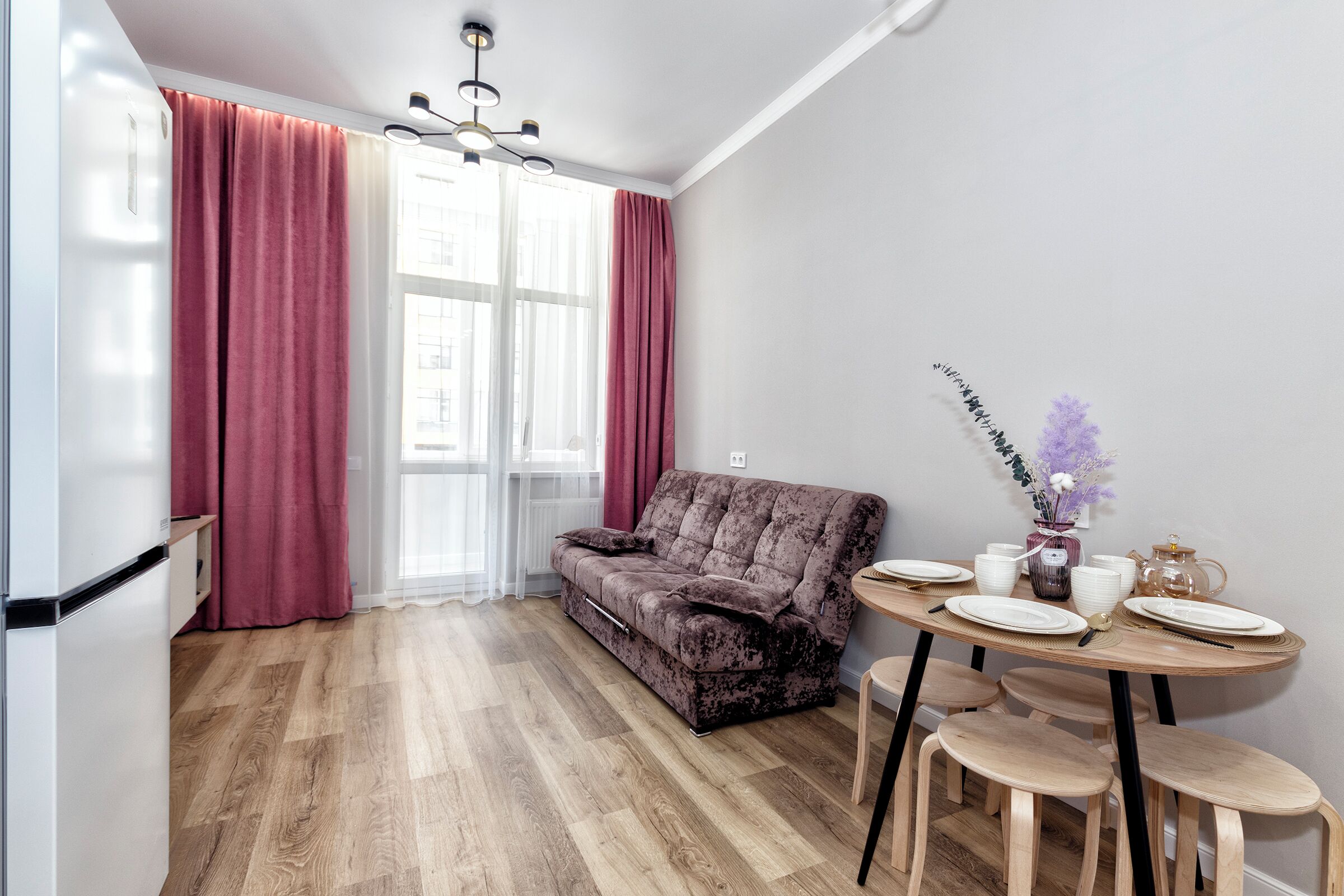 Un salon moderne avec un canapé, une table à manger et des chaises. On y trouve une fenêtre avec des rideaux, un lustre et un vase rempli de fleurs sur la table.