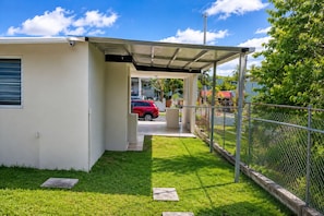 A covered outdoor area with a car parked inside, a fenced garden, and a building with a window and a door.