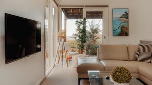 Een moderne woonkamer met een grote flatscreen televisie, een beige bank, een glazen salontafel en een schilderij van een kustlandschap.