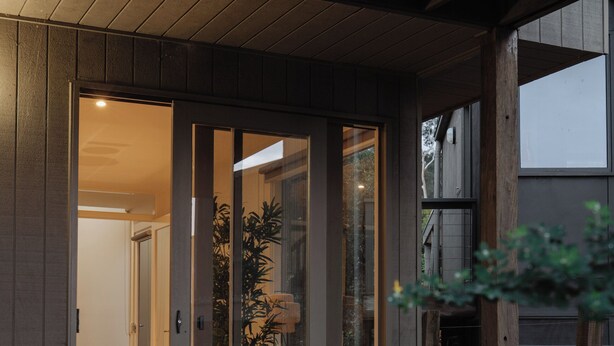 Een modern huis met een houten gevel, een glazen deur die leidt naar een goed verlicht interieur, en een kleine tuin met planten en een boomstronk.