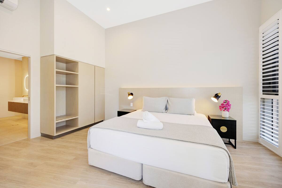 A modern bedroom with a large bed, built-in wardrobe, bedside lamps, and a window with blinds.