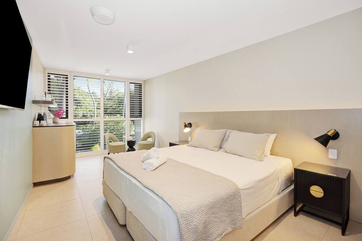 A modern bedroom with a large bed, a bedside table with a lamp, a small desk with a chair, and a window with blinds.