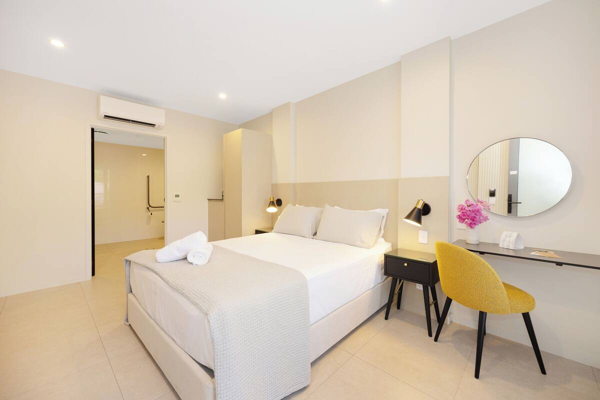 A hotel room with a bed, bedside table, chair, and a mirror on the wall.
