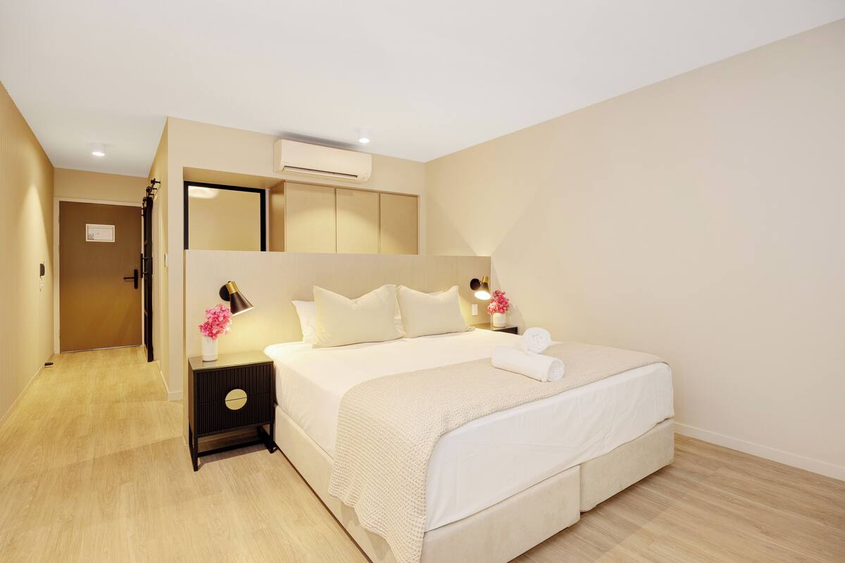 A bedroom with a bed, bedside tables, a nightstand, a mirror, and an air conditioning unit.