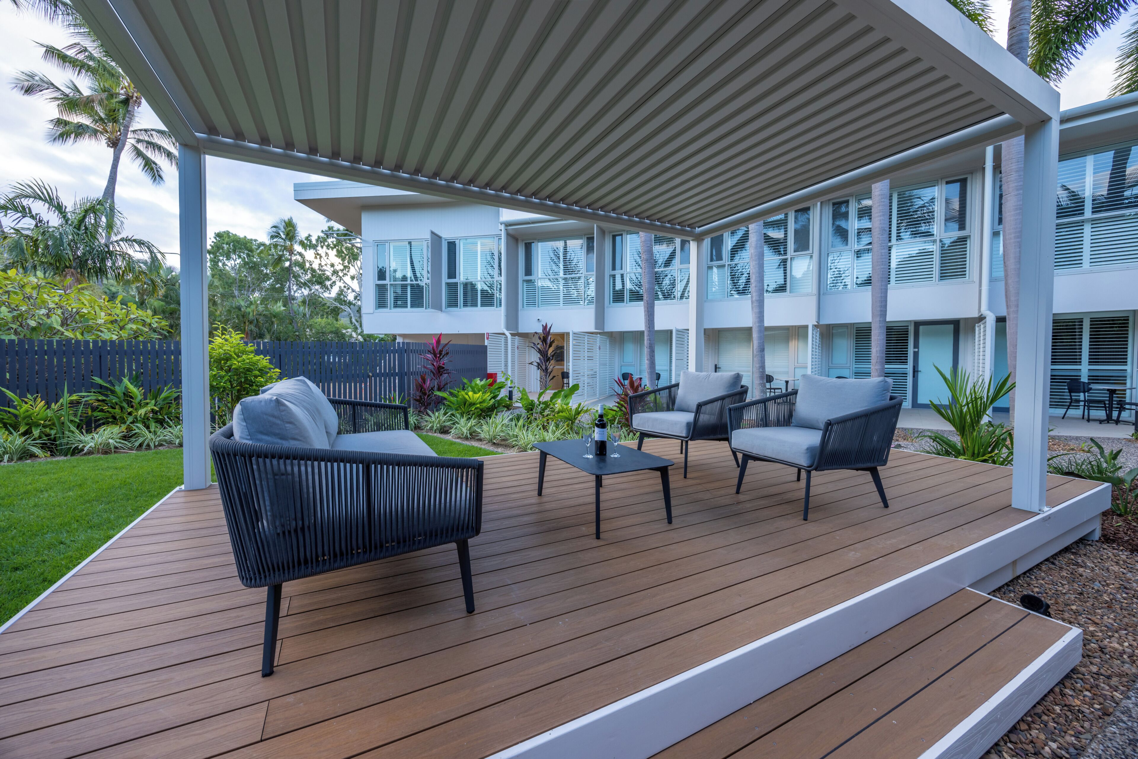 Un patio techado al aire libre con mobiliario moderno, una terraza de madera y vista a un edificio contemporáneo.
