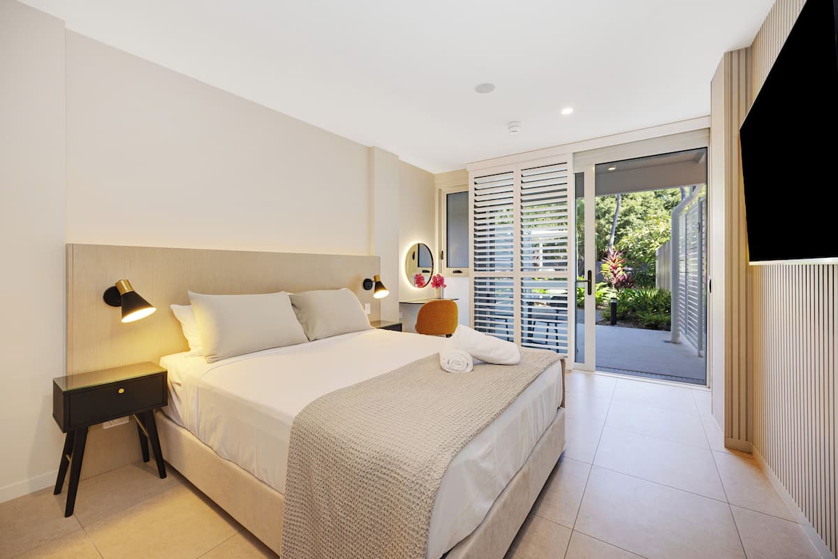A modern hotel room with a large bed, bedside table, lamp, and a view of the garden through the sliding doors.