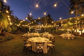 Un área de comedor al aire libre con mesas y sillas redondas, luces colgantes y palmeras al fondo.