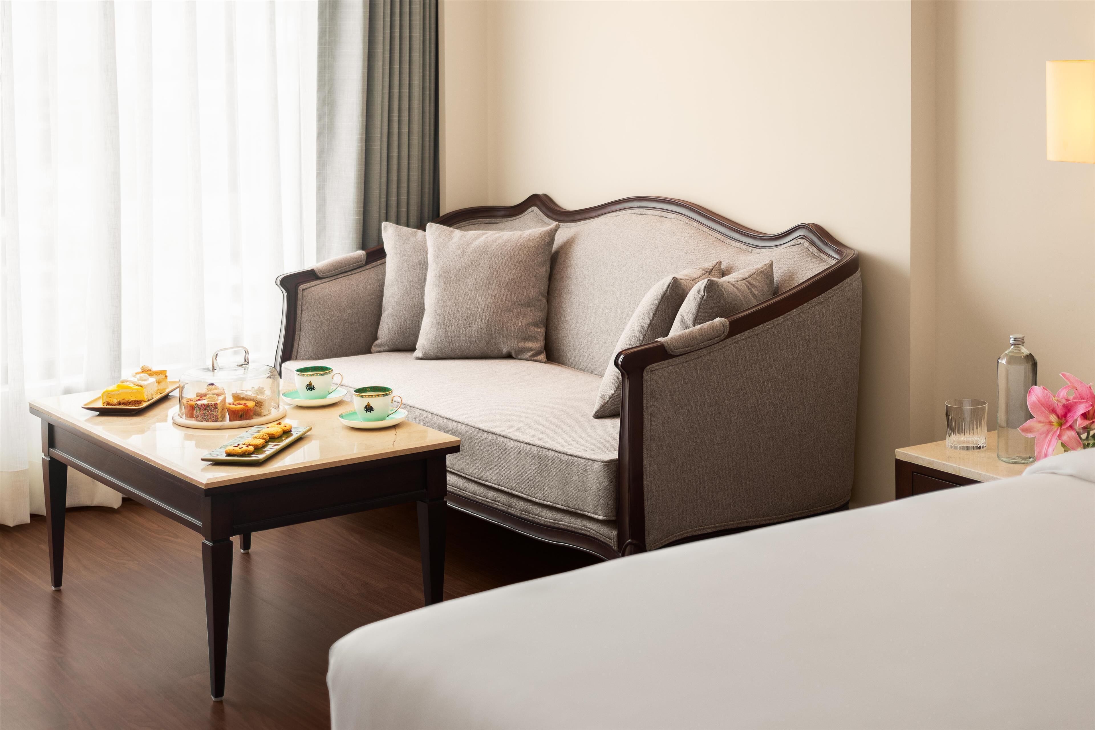 Habitación de hotel con sofá, una mesa de comedor con pasteles, un vaso de agua y un jarrón con flores.