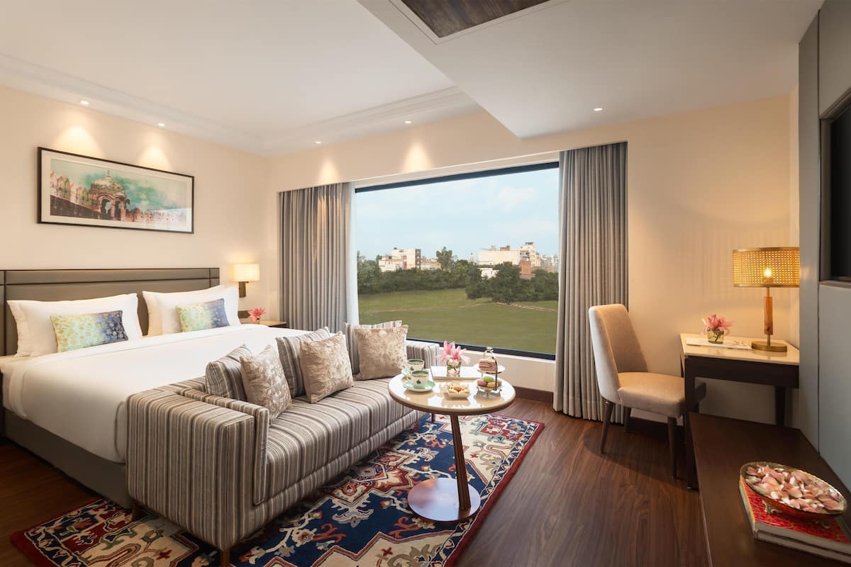 Une chambre d’hôtel avec un grand lit, un canapé, une petite table agrémentée d’objets décoratifs, un fauteuil et une vue sur le paysage urbain, visible à travers de grandes fenêtres.