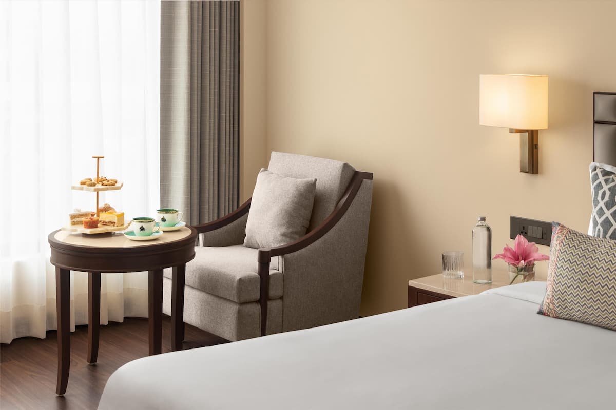 Une chambre d’hôtel avec un lit, une chaise, une petite table sur laquelle se trouve un plateau de nourriture, une lampe et un vase avec une fleur.