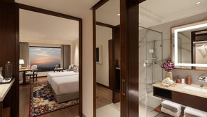 A hotel room with a bed, desk, and chair, a large window with a view, a bathroom with a mirror, sink, and towel rack.