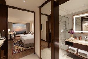Habitación de hotel con cama, escritorio y silla, un amplio ventanal con vistas, y baño con espejo, lavamanos y toallero.