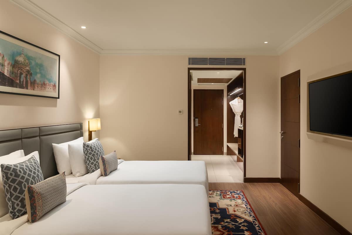 Une chambre d’hôtel avec deux lits, une télévision, un tableau et un couloir menant à un placard.