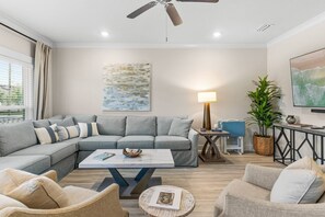 Un salon moderne comprenant un canapé d’angle, une table basse, une table d’appoint avec une lampe, un téléviseur et une grande fenêtre avec des rideaux.