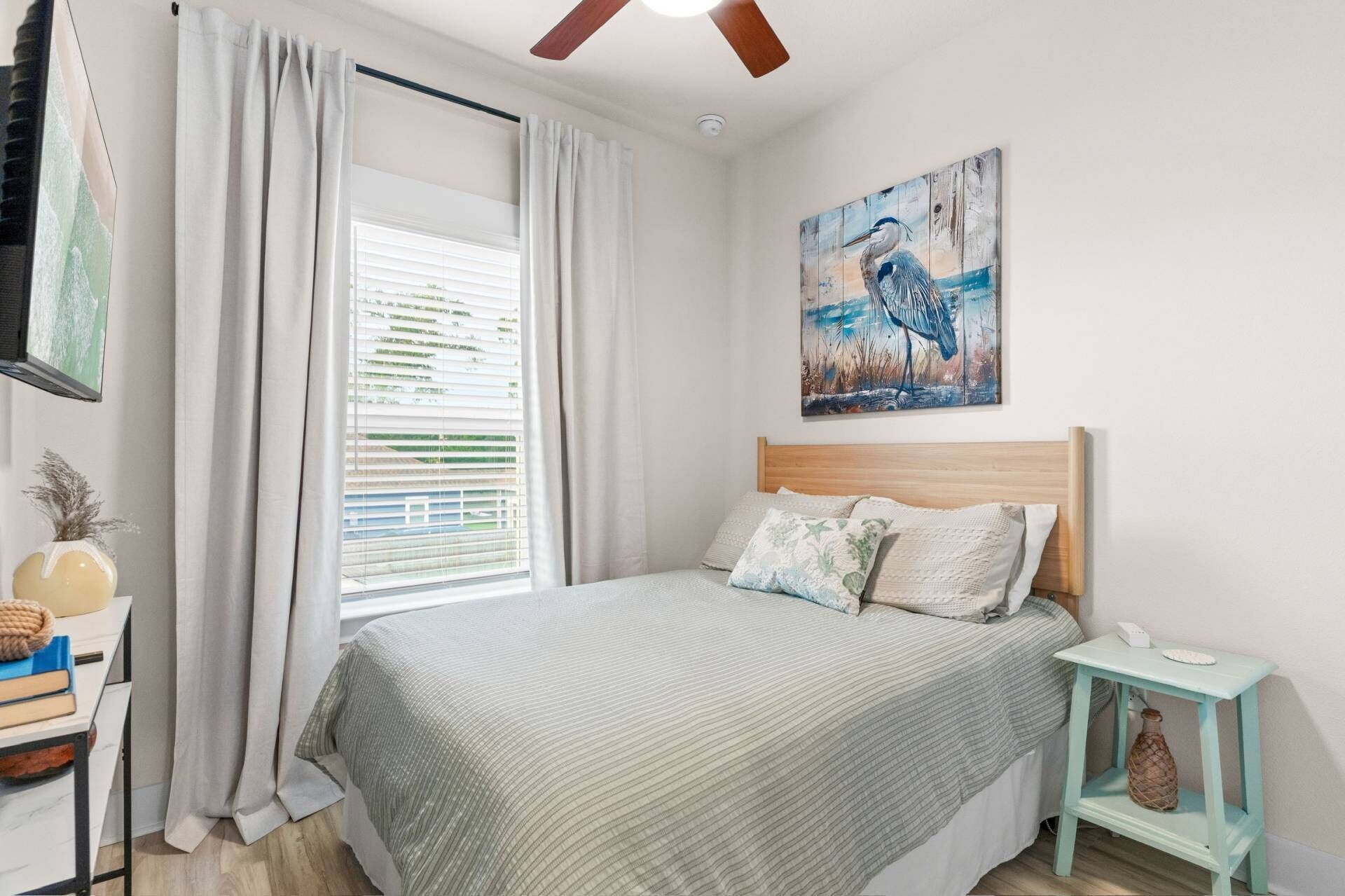 A bedroom with a bed, a window with curtains, a painting on the wall, a bedside table, and a ceiling fan.